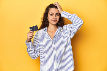 Woman holding a credit card, yellow backdrop, being shocked, she has remembered important meeting.