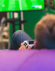 Process of playing video games, video game console with wireless controller, male kid child gamer on a couch bean bag chair, with black joystick in front of big wide screen tv, football video game © tsuguliev