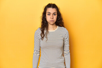 Young Caucasian woman, yellow studio background, blows cheeks, has tired expression. Facial expression concept. - obrazy, fototapety, plakaty