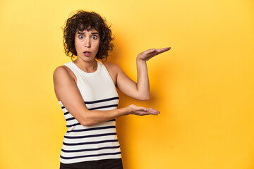 Caucasian curly-haired woman in white tank-top shocked and amazed holding a copy space between hands.