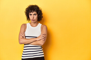 Caucasian curly-haired woman in white tank-top frowning face in displeasure, keeps arms folded.