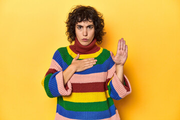 Curly-haired woman in multicolor sweatshirt taking an oath, putting hand on chest.