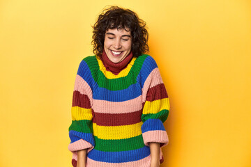 Curly-haired woman in multicolor sweatshirt laughs and closes eyes, feels relaxed and happy.