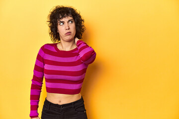 Caucasian curly-haired woman in pink striped-top touching back of head, thinking and making a choice.