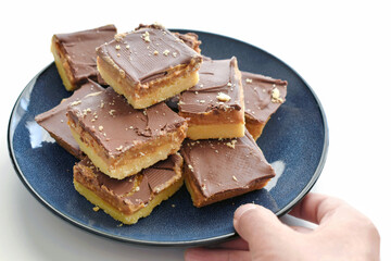 Millionaire's shortbread with chocolate and caramel on a blue plate. Hand serving chocolate caramel bars on the table.