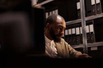 African american cop conducting investigation in police archive, sitting near shelf full of folder...