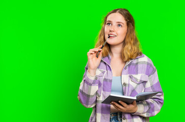 Thoughtful journalist young woman making notes, writing down thoughts with pen into notepad notebook diary, to do list, good idea. Pretty redhead student girl isolated on green chroma key background