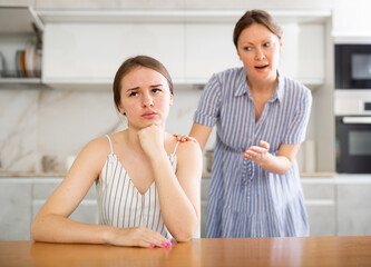 Upset teenage girl sitting at the kitchen-table turning her back upon mother attempting to come to terms with daughter
