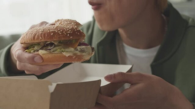 Medium closeup of delighted Asian boy with blond hair eating saucy big burger with pleasure at home