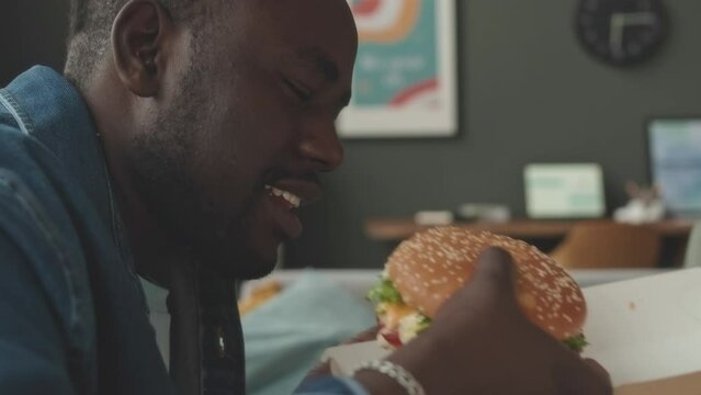 Tilt up of joyful young African American man eating delicious burger while having house party with friends on weekend