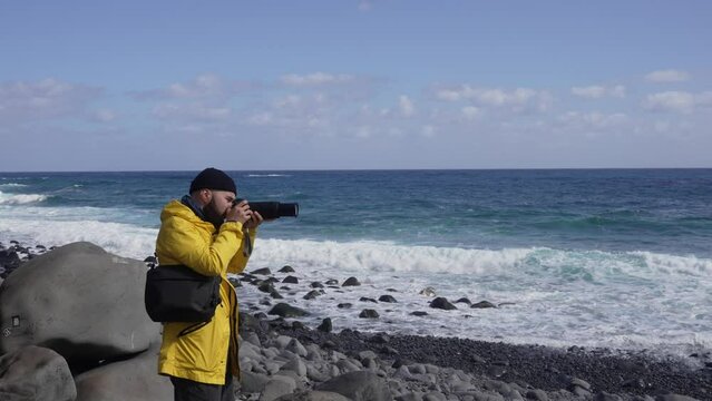 Man with a camera wearing a yellow raincoat taking photos of the Ruins of St. George, São Jorge beach at northeast coast
