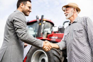 Investing in new tractor and agricultural equipment. Professional salesman in business suit shaking hands with senior farmer for successful purchase.