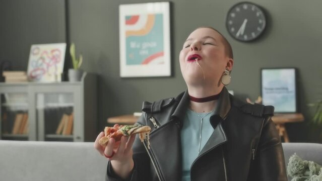 Medium shot of Caucasian plus size girl with shaved head looking at camera while eating delicious pizza sitting on couch in bright living room at daytimeMedium shot of Caucasian plus size girl with sh