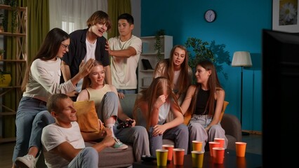 Group of teens, young people, friends sitting and standing around two guys, boys who were playing a console, gaming. One of them is celebrating his win emotionally. Full-size.