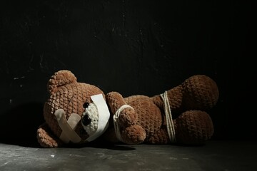Stop child abuse. Tied toy bear with taped mouth and patches lying on grey floor against black...