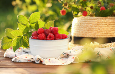 Tasty ripe raspberries in bowl, green leaves and straw hat on wooden table outdoors