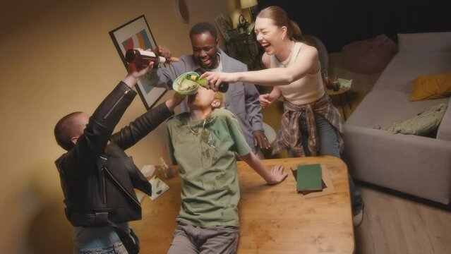 High angle shot of carefree young diverse people having party with alcohol at home, pouring beer into plastic funnel in Asian boys mouth