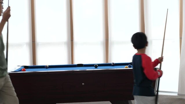 Asian child boy playing billiard or snooker with father. Kid concentrate on cue stick to strike cue ball to pot other balls to pocket. Concept of family sport game, leaning, leisure, goal.