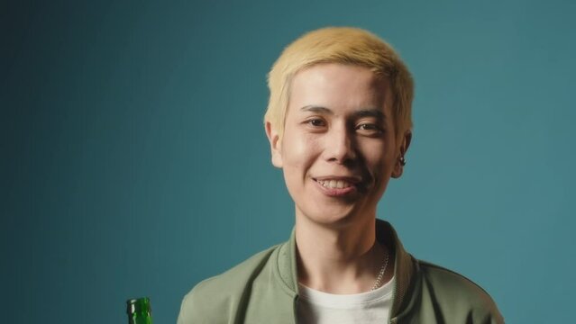 Chest up portrait of Asian guy with dyed blond hair posing for camera with bottle of non alcoholic beer standing on blue studio background