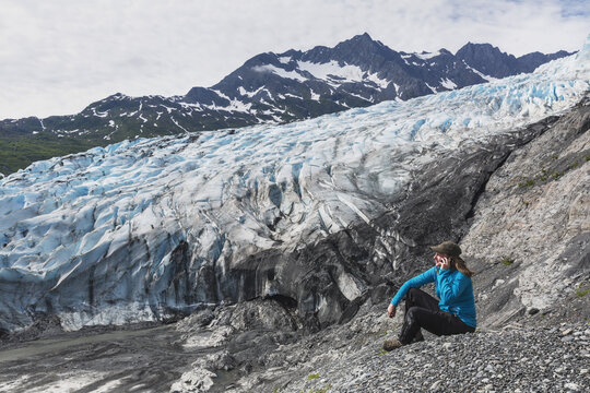 Woman Sitting On Overlook Talking On A Cell Phone In Front Of Shoup Glacier, Shoup Bay State Marine Park, Prince William Sound; Valdez, Alaska, United States Of America