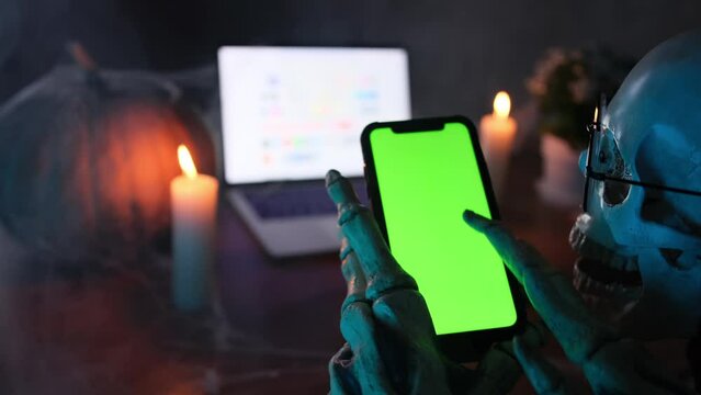 Hands of skeleton holding smartphone with chromakey scrolls social networks, scull looks screen, in cloud of smoke, vignette. Halloween festive atmosphere. Communicating on social networks. 