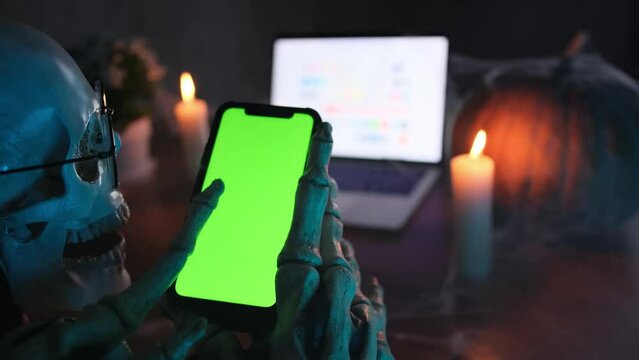 In fog hands in skeleton costume touch smartphone scrolls green screen, mystical dark atmosphere, burning candles flicker mysteriously on blurred background with laptop. Creepy Halloween. 