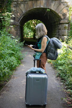 Mature woman uses her smart phone while standing on a path with her luggage; United Kingdom