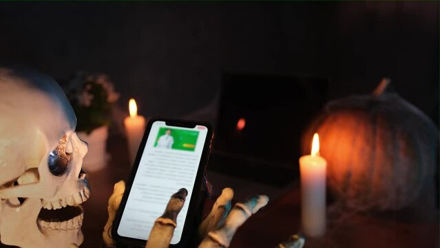 Funny scary skeleton holds a phone with bones hands, reads articles and creepy posts on sites on internet in puffs of smoke on candlelight in festive gloomy atmosphere of Happy Halloween.