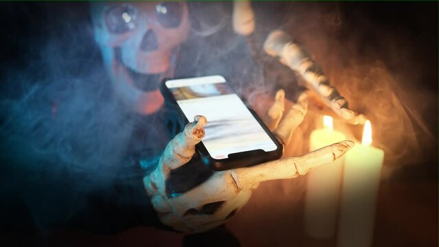 Сreepy background, invitation to Halloween holiday with skeleton scrolling through social networks on smartphone screen, light of burning candles and flowing fog on dark background, vignette.