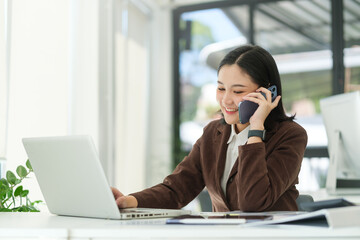 Cheerful asian female employs sitting front of her laptop and talking on mobile phone.