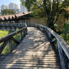Obraz premium Small wooden bridge surrounded by trees with small chalets inside the woods. Modern buildings in the background