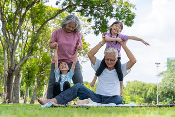 Grandparents let their granddaughter sit on their shoulder in the park. People of different generations laugh happily in the park on the mats.