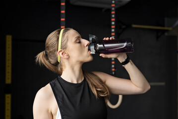 Athletic woman drinking water after physical workout in a gym on dark background. 