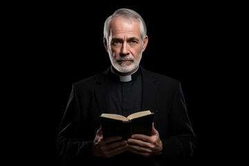 Catholic christian church priest wearing black cassock robe holding the holy bible book in his hands. Isolated on dark black background.