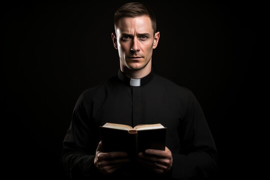 Catholic christian church priest wearing black cassock robe holding the holy bible book in his hands. Isolated on dark black background.