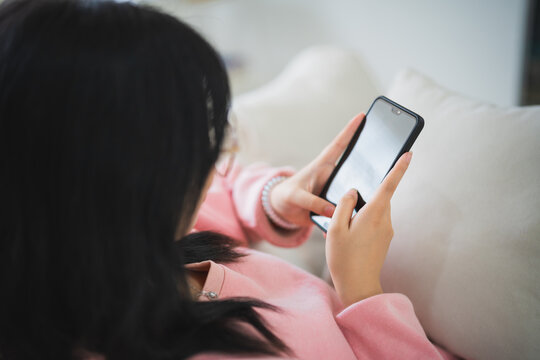 Smiling asian woman wearing casual clothes sitting on a couch sofa using mobile phone to social media or seach jobs or chatting or trade stock at the living room.