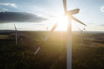 Wind turbines in a hilly forest in front of a partly cloudy, but sunny sky are seen from an aerial view during sunset