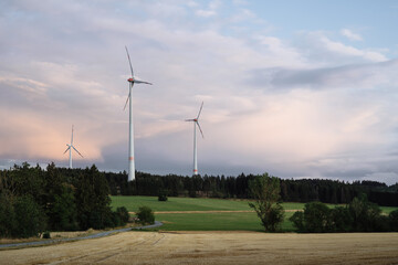 Fototapeta premium Wind turbines between trees in a forest in front of a beautifiul, pastel colored evening sky