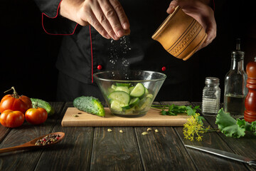 The chef prepares a vegetable salad in the restaurant kitchen. Adding salt to vitamin salad with cucumbers and tomatoes
