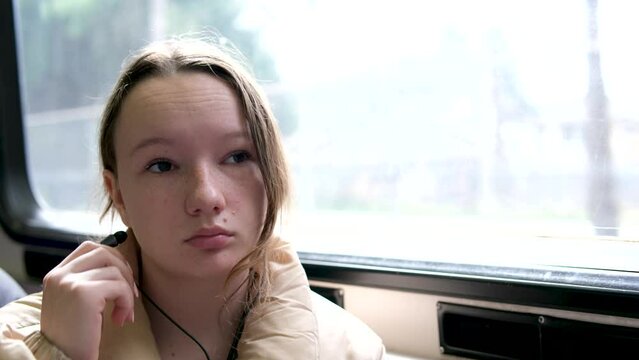 teenage girl rides in skytrain on train in bus headphones in ears listens to music autumn spring winter warm beige jacket hair is tied down real person sits tucked legs under sad dreams awaits stop