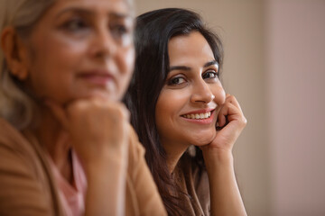 A YOUNG WOMAN SITTING AND SMILING WITH MOTHER SITTING NEAR AND THINKING