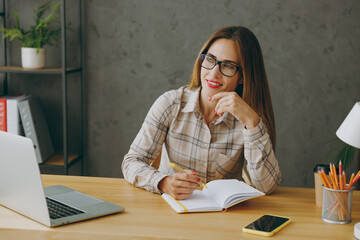 Young minded successful employee business woman wear shirt casual clothes glasses sit work at office desk with pc laptop, look aside write down in notebook, take notes. Achievement career job concept.