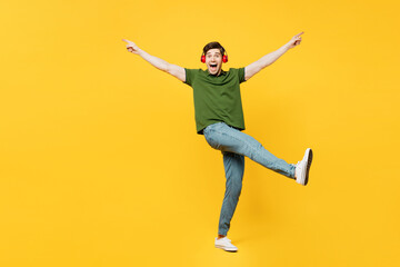 Fototapeta na wymiar Full body side view young happy man he wears green t-shirt casual clothes listen to music in headphones raise up hands leg dance isolated on plain yellow background studio portrait. Lifestyle concept.