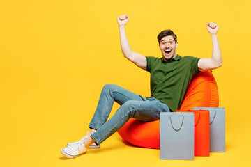Young overjoyed excited fun man wears casual clothes sit in bag chair near paper package bags after shopping do winner gesture isolated on plain yellow background. Black Friday sale buy day concept.