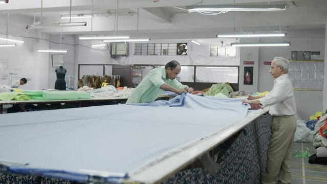 Two skilled Indian men or male employees are working with a big piece of cotton cloth or fabric in an indoor textile workshop or small-scale garment industry or factory