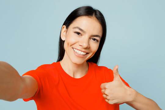 Close up young latin woman wear orange red t-shirt casual clothes doing selfie shot pov on mobile cell phone show thumb up isolated on plain pastel light blue cyan background studio Lifestyle concept