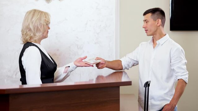 Young man handing over passport to find reserved room in hotel. Blonde female receptionist checking visitor reservation in computer