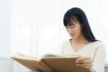Fototapeta premium Happy - cheerful Asian young woman with eye glasses sitting on the sofa in living room and reading a book alone.