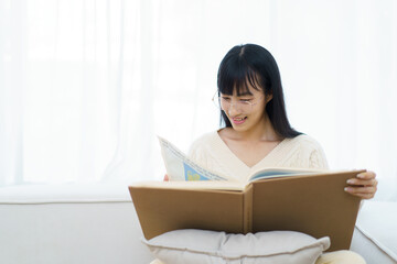 Happy - cheerful Asian young woman with eye glasses sitting on the sofa in living room and reading a book alone.