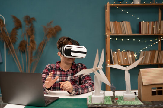 Schoolboy at home studying alternative energy wears virtual reality glasses while sitting at table with school project
